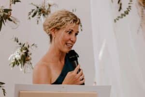 Eine Frau mit lockigem blondem Haar spricht bei einer Hochzeit in ein Mikrofon. Sie trägt ein One-Shoulder-Top und steht vor einem wunderschön arrangierten Hintergrund, der mit hängendem Grün und weißem Stoff dekoriert ist und die Essenz eleganter Hochzeitsplanung perfekt einfängt.