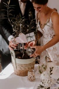 Braut und Bräutigam, elegant für ihre Hochzeit gekleidet, nehmen an einer Zeremonie zum Pflanzen eines Einheitsbaums teil. Sie füllen vorsichtig Erde aus Glasgefäßen in einen Topfbaum in einem dekorativen Korb, was Wachstum und Einheit symbolisiert. In der Nähe stehen Karten und eine kleine Gießkanne auf dem Tisch bereit.