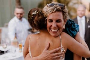Zwei Frauen umarmen sich herzlich bei einer Hochzeit im Freien. Eine hat ihr Haar zu einem mit Blumen geschmückten Knoten hochgesteckt, während die andere lockiges blondes Haar und eine Sonnenbrille auf dem Kopf hat. Sie lächeln beide, sind von Gästen in Abendgarderobe umgeben und feiern diesen freudigen Anlass.