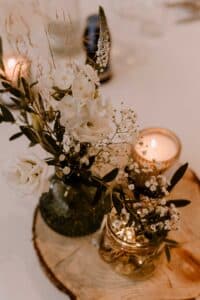 Ein rustikales Tischdekor, perfekt für eine Hochzeit, mit weißen Blumen und Grünpflanzen in Glasgefäßen auf einer Holzplatte. Eine brennende Kerze in einem kleinen Kerzenhalter verleiht dem bezaubernden Arrangement einen warmen Schein, ideal für jede Hochzeitsplanung.