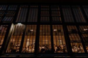 Durch große Fenster fällt der Blick in einen warm beleuchteten Raum, in dem Menschen bei einer Feier zusammensitzen, vielleicht in einer Hochzeitslocation. Die Atmosphäre ist lebhaft und gesellig, die Tische sind elegant für den freudigen Anlass einer Hochzeit gedeckt.