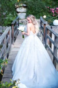 Eine Braut in einem trägerlosen weißen Kleid hält einen Strauß weißer Blumen in der Hand, während sie auf einer Holzbrücke steht. Sie blickt über ihre Schulter zurück, umgeben von üppigem Grün und rosa Blumen – und fängt die Magie eines perfekten Hochzeitsmoments ein.