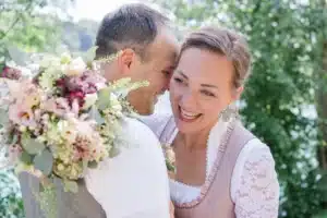 Ein Paar in traditioneller Kleidung lächelt fröhlich in einer üppigen Umgebung im Freien und feiert seine Hochzeit. Der Mann hält einen Strauß bunter Blumen und die Frau lehnt sich an ihn. Beide wirken glücklich und entspannt mit Bäumen und Grün im Hintergrund.