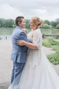 Ein Paar im Hochzeitskleid umarmt sich draußen an einem See und fängt die Essenz ihrer Hochzeit ein. Der Bräutigam trägt einen hellblauen Anzug, während die Braut in einem weißen Kleid mit Spitzendetails und einem transparenten Schal glänzt. Sie stehen auf einem Kiesweg, umgeben von Grün und Bäumen unter einem bewölkten Himmel.
