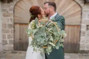 Ein Paar küsst sich vor einer rustikalen Holztür, eine Erinnerung an ihre sorgfältige Hochzeitsplanung. Sie halten ein kreisförmiges Blumenarrangement mit Eukalyptuszweigen hoch, das ihre Gesichter teilweise verdeckt. Die Frau trägt ein weißes Kleid und der Mann einen grünen Anzug, verloren in der Romantik des Heiratens.