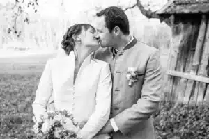Ein Schwarzweißfoto zeigt ein Paar, das sich während seiner Hochzeit im Freien küsst. Die Frau hält einen Blumenstrauß, beide in traditioneller Kleidung stehen neben einer rustikalen Holzkonstruktion, eingerahmt von Bäumen. Dieser zeitlose Moment verkörpert die Essenz romantischer Hochzeitsfeiern.