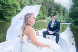 Eine Braut in einem weißen Kleid hält einen weißen Regenschirm und sitzt neben einem Bräutigam in einem schwarzen Anzug mit einer grünen Ansteckblume. Sie befinden sich in einem kleinen Boot auf einem ruhigen See, umgeben von üppigen grünen Bäumen – eine heitere und malerische Hochzeitsszene, wie aus einem Heiratstraum.