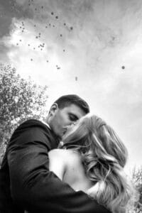 Ein Schwarzweißfoto zeigt ein sich küssendes Paar unter freiem Himmel, das ihre Reise zur Hochzeit symbolisiert, über dem Luftballons schweben. Der Mann trägt einen Anzug und die Frau hat langes, welliges Haar. Im Hintergrund dieses bezaubernden Hochzeitsplanungsmoments stehen majestätisch Bäume.