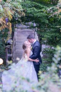 Ein Paar im Hochzeitskleid küsst sich vor einer rustikalen Holzbrücke, umgeben von üppigem Grün, und verkörpert damit die Freude am Heiraten. Die Braut trägt ein fließendes weißes Kleid und der Bräutigam einen dunklen Anzug. Diese romantische Szene fängt die Essenz einer Hochzeit im Freien perfekt ein.
