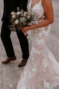Eine Braut in einem geblümten Spitzenbrautkleid hält einen Blumenstrauß in der Hand, während sie neben einer Person im dunklen Anzug auf einem Sandweg geht und so die perfekte Szene für ihre Hochzeit schafft.