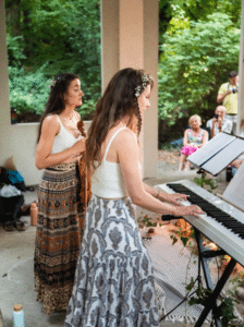 Zwei Frauen, beide in langen Röcken und weißen Oberteilen, spielen im Freien bei einer Hochzeit bezaubernde Musik. Eine spielt Geige, während die andere in einem geschützten Bereich, umgeben von üppigen Pflanzen, die Klaviatur ziert und die sitzenden Gäste begeistert, die an der fröhlichen Hochzeitsplanung beteiligt sind.