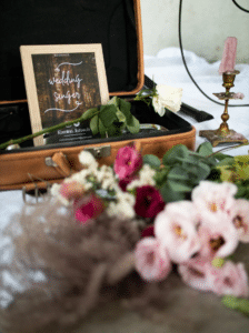 Auf einem Vintage-Koffer befindet sich ein gerahmtes Schild mit der Aufschrift „Hochzeitssängerin“ und einer weißen Rose daneben, perfekt für Ihre Hochzeit. Im Vordergrund sind verschwommene rosa und rote Blumen neben einem Kerzenständer mit einer rosa Kerze angeordnet. Die Szene ist auf einem weißen Tuch arrangiert, ideal für die Hochzeitsplanung.