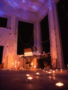Ein gemütliches, stimmungsvolles Ambiente für eine romantische Hochzeit, mit einer Person, die in einem schwach beleuchteten Raum Keyboard spielt. Lichterketten bedecken die Wände und Kerzen beleuchten den Boden. Eine Sternenlichtprojektion wirft sanfte Blautöne an die Decke und verstärkt diese heitere Hochzeitsatmosphäre.