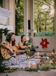 Zwei Menschen sitzen mit Gitarren auf einer Veranda, umgeben von Pflanzen, Kerzen und Blumen. Sie lächeln in dieser entspannten Umgebung, die sich perfekt für eine intime Hochzeitsplanung eignet, mit einem Wald als Hintergrund. Auf die Wand hinter ihnen ist ein roter Stern gemalt.