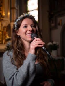 Eine Frau mit langen braunen Haaren, einem grauen Pullover und einem geblümten Stirnband, lächelt und hält ein Mikrofon. Sie steht drinnen, wahrscheinlich in einer Kirche – die perfekte Kulisse für Hochzeitsplanung – mit verschwommenen Blumendekorationen und Buntglas im Hintergrund.
