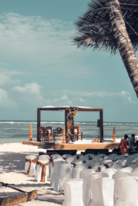 Strandhochzeits-Setup mit weißen Stühlen mit goldenen Schärpen vor einem mit Blumenschmuck geschmückten Holzaltar – perfekt für alle, die ihre Traumhochzeit planen. Der Veranstaltungsort ist von Palmen beschattet, im Hintergrund das Meer und ein teilweise bewölkter Himmel.