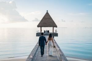 Ein Paar geht Hand in Hand über einen Holzsteg zu einem strohgedeckten Pavillon über ruhigem, blauem Wasser, perfekt für eine Hochzeit. Der Himmel ist klar mit sanften Wolken, was eine ruhige und romantische Atmosphäre schafft, ideal für die Hochzeitsplanung.