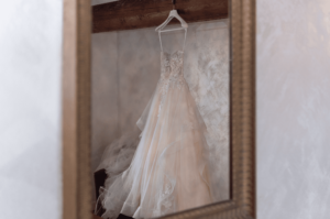 Ein Spitzenbrautkleid, perfekt für die Hochzeitsplanung, hängt elegant auf einem Bügel und spiegelt sich in einem rustikalen Spiegel mit Holzrahmen. Das Kleid zeigt komplizierte Blumenmuster und einen mehrlagigen Tüllrock und weckt ein Gefühl von Romantik und Vorfreude auf die bevorstehende Hochzeit.