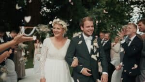 Braut und Bräutigam lächeln, als sie bei ihrer Hochzeit im Freien den Mittelgang entlanggehen, umgeben von Gästen, die Blütenblätter werfen. Die Braut trägt ein weißes Kleid mit einem Blumenkranz und der Bräutigam einen Anzug mit Fliege, umrahmt von der üppigen, grünen Umgebung.