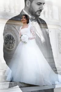 Eine Braut in einem Spitzenhochzeitskleid steht anmutig da und hält einen Blumenstrauß in der Hand. Der durchscheinende Überwurf des Bräutigams im Anzug ist sichtbar und fängt die Essenz von Hochzeit und Romantik ein. Eine Treppe mit kunstvollen Details dient als Kulisse für diesen bezaubernden Moment.