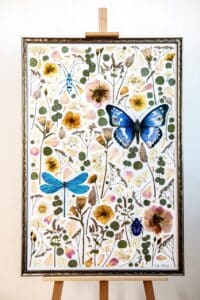 Ein gerahmtes Kunstwerk auf einer Staffelei, perfekt als Hochzeitsdekoration, mit detaillierten Abbildungen von Schmetterlingen, Libellen und verschiedenen Blumen und Pflanzen vor einem weißen Hintergrund. Die lebendigen Farben umfassen Blau-, Gelb- und Grüntöne und verleihen jeder Hochzeitsplanung eine lebendige Note.