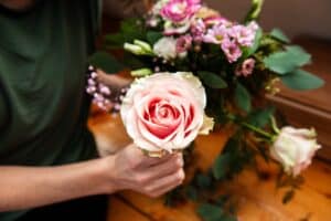 Eine Person in einem grünen Hemd arrangiert sorgfältig einen Blumenstrauß auf einer Holzoberfläche und perfektioniert damit das Blumenarrangement für eine elegante Hochzeit. Die rosa Rose liegt sanft neben rosa und weißen Blüten und fängt die Essenz zeitloser Liebe und sorgfältiger Hochzeitsplanung ein.