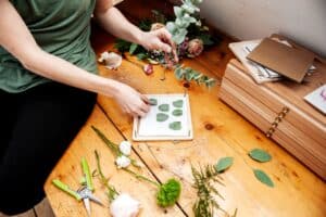Eine Person arrangiert Eukalyptusblätter in einer Holzpresse auf einem Tisch und bereitet damit die Bühne für eine elegante Hochzeit. In der Nähe liegen Blumen, eine grüne Schere und gestapelte Holzkisten, die alle die Kunst des Blumenarrangierens und -bastelns als Teil einer sorgfältigen Hochzeitsplanung betonen.