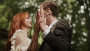 Ein Paar in Hochzeitskleidung steht sich in einem Wald gegenüber und verkörpert einen ruhigen Moment der Hochzeitsplanung. Ihre sich sanft berührenden Handflächen stehen im Mittelpunkt, wobei die roten Haare und Tattoos der Frau den Anzug des Mannes vor einem verschwommenen Hintergrund aus Grün ergänzen.