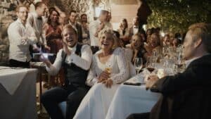 Ein fröhliches Paar sitzt bei seinem Hochzeitsempfang an einem Tisch, umgeben von klatschenden und lächelnden Gästen. Der Veranstaltungsort ist warm beleuchtet, mit rustikalen Steinmauern und grünem Laub. Weingläser und Dekorationen schmücken die Tische und spiegeln eine tadellose Hochzeitsplanung wider.