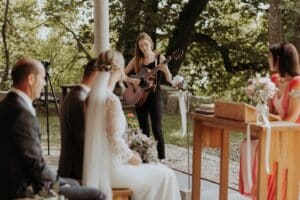Eine Frau klimpert auf einer Akustikgitarre und untermalt die intime Hochzeitszeremonie mit gefühlvollen Melodien. Braut und Bräutigam hören aufmerksam zu, umgeben von Gästen inmitten üppiger Vegetation und rustikaler Holzmöbel – ein exquisiter Moment inmitten ihrer sorgfältig geplanten Hochzeit.