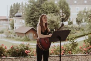 Eine Frau steht im Freien, spielt Gitarre und singt in ein Mikrofon. Sie ist von einem malerischen Garten mit blühenden Blumen und Bäumen umgeben – perfekt als Hochzeitskulisse. Im Hintergrund ist ein Gebäude zu sehen, das zur bezaubernden Atmosphäre dieses besonderen Tages beiträgt.