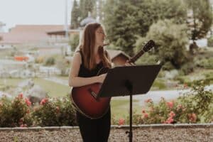 Eine Frau mit langen Haaren steht draußen, spielt Gitarre und singt, vielleicht um einem Paar an seinem besonderen Tag ein Ständchen zu bringen. Sie steht neben einem Notenständer, umgeben von blühenden roten Blumen und der üppigen Kulisse eines Hochzeitsgartens in voller Blüte, perfekt für jede Hochzeitsplanung.