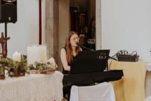 Ein Musiker mit langen Haaren und Brille spielt Keyboard und singt in ein Mikrofon und schafft so die Stimmung einer romantischen Hochzeit. Die Szene umfasst Kerzen und ein Blumenarrangement auf einem Tisch im Vordergrund und suggeriert eine ruhige Umgebung, die perfekt für jede Hochzeitsplanung ist.