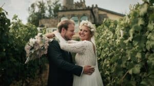 Braut und Bräutigam umarmen sich freudig in einem Weinberg, umgeben von üppigen grünen Weinreben. Die Braut, die eine Blumenkrone und ein Spitzenkleid trägt, hält einen Blumenstrauß, während der Bräutigam einen dunklen Anzug trägt. Diese malerische Kulisse ist perfekt für diejenigen, die ihre Traumhochzeit planen und dabei jedes Detail inmitten des Charmes der Geschichte planen.