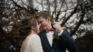 Während ihrer Hochzeitsplanung teilen Braut und Bräutigam einen freudigen Moment unter einem durchsichtigen Regenschirm. Der Bräutigam trägt einen marineblauen Anzug mit roter Fliege und Boutonniere, während die Braut im Spitzenkleid vor kahlen Bäumen steht und eine rustikale Kulisse schafft, die die romantische Atmosphäre verstärkt.