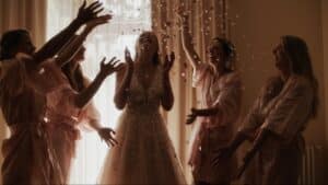 Eine Braut im Brautkleid lächelt freudig, während sie ihre Hochzeit feiert, während vier Brautjungfern in passenden Gewändern Blütenblätter in die Luft werfen. Der warm beleuchtete Raum verstärkt diesen Moment der Hochzeitsplanung mit einer festlichen und feierlichen Atmosphäre.