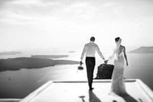 Ein Schwarzweißfoto von Braut und Bräutigam, die Händchen haltend auf einen malerischen Meerblick zulaufen, fängt die Essenz ihrer Hochzeit ein. Die Braut trägt ein fließendes Kleid und einen Schleier, während der Bräutigam Hemd und Hose trägt. Im Hintergrund erstrecken sich Inseln und das Meer und verkörpern zeitlose Romantik.