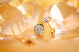 Nahaufnahme von mit Diamanten besetzten goldenen Ohrringen, die auf einer reflektierenden Oberfläche ruhen, perfekt für jede Hochzeit. Daneben hängt eine zarte Goldkette, die mit ihren glänzenden Gold- und Diamantakzenten eine warme und elegante Komposition schafft, ideal für diejenigen, die gerade mitten in der Hochzeitsplanung stecken.