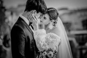 Auf einem zeitlosen Schwarzweißfoto teilen Braut und Bräutigam einen intimen Moment, umarmen sich während ihrer Hochzeit. Die Braut in ihrem Spitzenkleid und Schleier berührt zärtlich das Gesicht des Bräutigams, während sie einen Blumenstrauß hält, und fängt so einen kostbaren Moment der Liebe vor einem sanften Hintergrund ein.