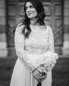 Eine Frau steht vor einer detailreichen Holztür. Sie trägt ein langärmliges Kleid mit Blumenmuster. Sie hält einen Strauß Rosen in der Hand und hat langes, welliges Haar. Das Bild fängt die Essenz von heiraten in zeitloser schwarz-weißer Eleganz ein.