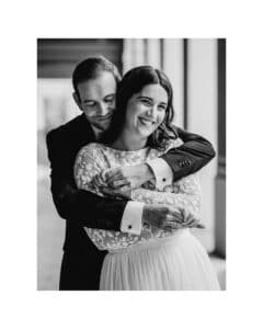 Ein lächelndes Paar umarmt sich herzlich, wobei der Mann seine Arme von hinten um die Frau legt. Sie trägt ein weißes geblümtes Kleid und er einen Anzug, der die Freude einer Hochzeit verkörpert. Die Inneneinrichtung ist sanft beleuchtet und verstärkt das zeitlose Gefühl dieses schwarz-weißen Moments.