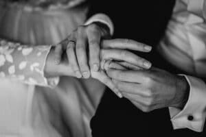 Ein Nahaufnahmefoto in Schwarzweiß fängt den intimen Moment eines Ringtauschs während einer Hochzeit ein. Eine Person steckt der anderen zärtlich den Ring an den Finger, beide tragen formelle Kleidung, wobei an einem Ärmel ein zartes Stoffdetail zu sehen ist, das die Schönheit des Heiratens hervorhebt.