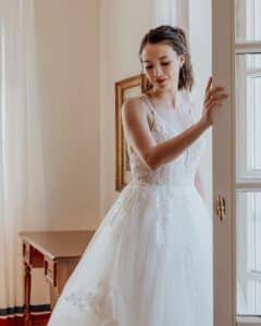 Eine Frau in einem weißen Spitzenbrautkleid steht neben einem Fenster und hält sanft den Türrahmen. Der Raum, perfekt für eine intime Hochzeitsplanung, hat helle Wände und Vorhänge, mit einem Holztisch im Hintergrund.