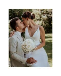 Braut und Bräutigam stehen in einem Garten, eingefangen in einem romantischen Kuss, der die Essenz einer Hochzeit verkörpert. Sie trägt ein weißes Kleid, ihr Strauß weißer Rosen passt zu seinem hellen Anzug, alles vor einem Hintergrund üppiger Vegetation.
