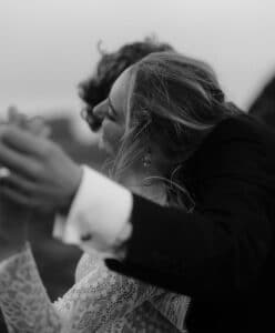 Schwarz-weiße Nahaufnahme eines Paares, das vielleicht mitten in seiner Hochzeit steht und im Freien tanzt. Die Frau im Spitzenkleid lehnt sich leicht zurück und hält die Hand des Mannes. Beide wirken fröhlich und entspannt, die unscharfe Umgebung lässt Bewegung vermuten.
