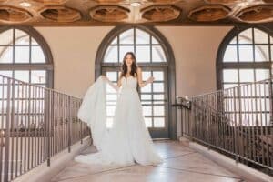 Eine lächelnde Braut steht auf einem Balkon mit verziertem Geländer und genießt den Moment ihrer Hochzeit, während sie ihr fließendes weißes Kleid hält. Die Holzdecke darüber sorgt für Wärme, während große Bogenfenster natürliches Licht hereinlassen und den bezaubernden Raum erhellen.
