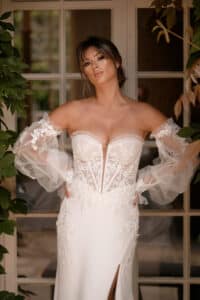 Eine Frau in einem eleganten weißen Hochzeitskleid mit floralen Spitzendetails und transparenten schulterfreien Ärmeln steht vor einer Glastür, umgeben von üppigen grünen Blättern, und verkörpert den perfekten Moment in jeder Hochzeitsplanungsvision.