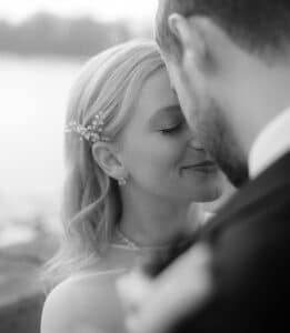 Eine Nahaufnahme eines Paares in Schwarzweiß, das sich im Freien umarmt und einen liebgewonnenen Auftakt zu ihrer Hochzeit einfängt. Die Frau, die die Augen geschlossen hat und ein Haar-Accessoire trägt, lehnt sich in den Moment hinein, während sich der Mann leicht abwendet, alles eingerahmt von der ruhigen Kulisse eines entfernten Gewässers.