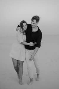 Ein Schwarzweißfoto von einem Mann und einer Frau, die barfuß an einem Sandstrand spazieren gehen, fängt die Freude ihrer Hochzeit ein. Die Frau in einem hellen Kleid und der Mann in einem dunklen Hemd und hellen Hosen lächeln herzlich, als sie sich auf dieser unvergesslichen Reise nach Heiraten umarmen.