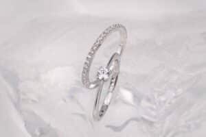 Zwei elegante Ringe auf einer reflektierenden Oberfläche: Einer ist ein Solitärring mit Diamanten, der andere ist ein mit kleinen, funkelnden Steinen verzierter Ring. Beide sind in glattes, silbriges Metall eingefasst. Der sanft verschwommene Hintergrund verstärkt ihren Glanz – eine perfekte Wahl für alle, die sich intensiv mit der Hochzeitsplanung beschäftigen.